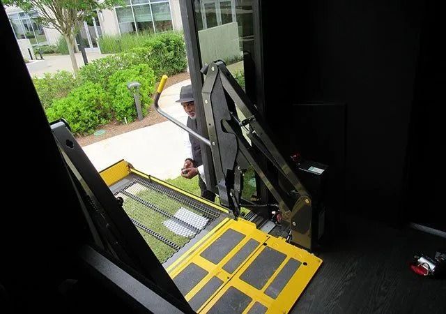 A wheelchair lift partially deployed from a vehicle, with a person standing on the sidewalk waiting nearby.