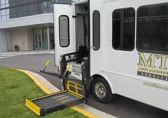 A white shuttle bus parked on a curb with its mechanical wheelchair lift extended and lowered to the ground.