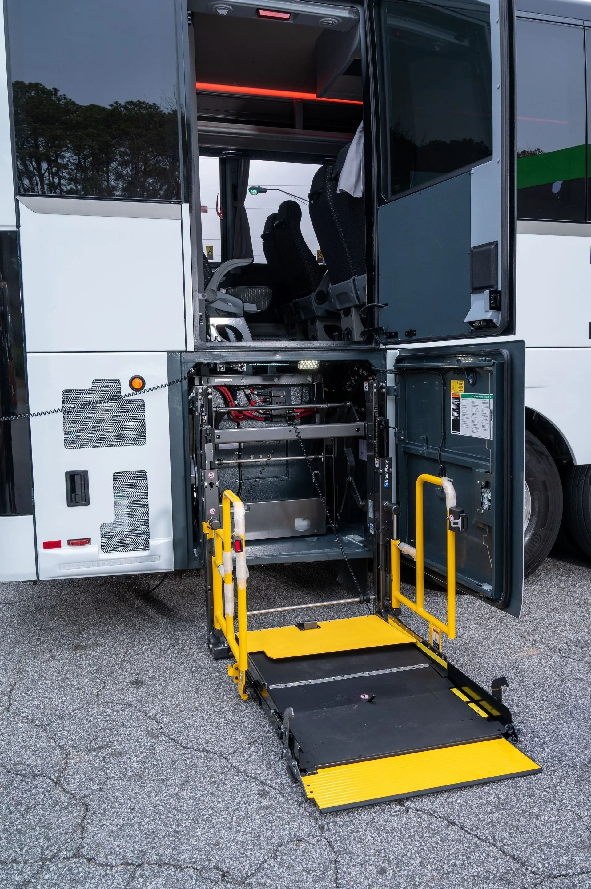 A white bus with an extended yellow wheelchair lift deployed at the side entrance, parked on a gravel surface.