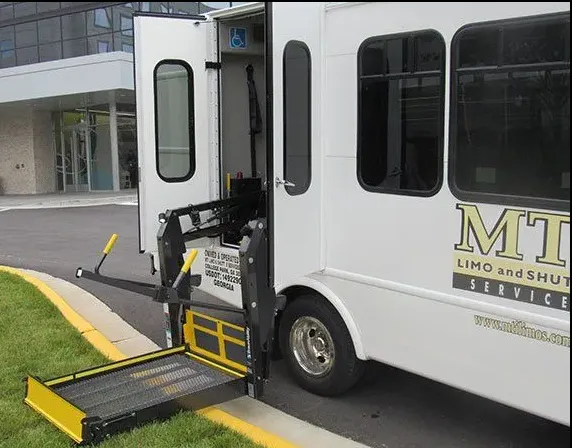 A white shuttle bus with an extended mechanical wheelchair lift deployed at the curb in front of a building.