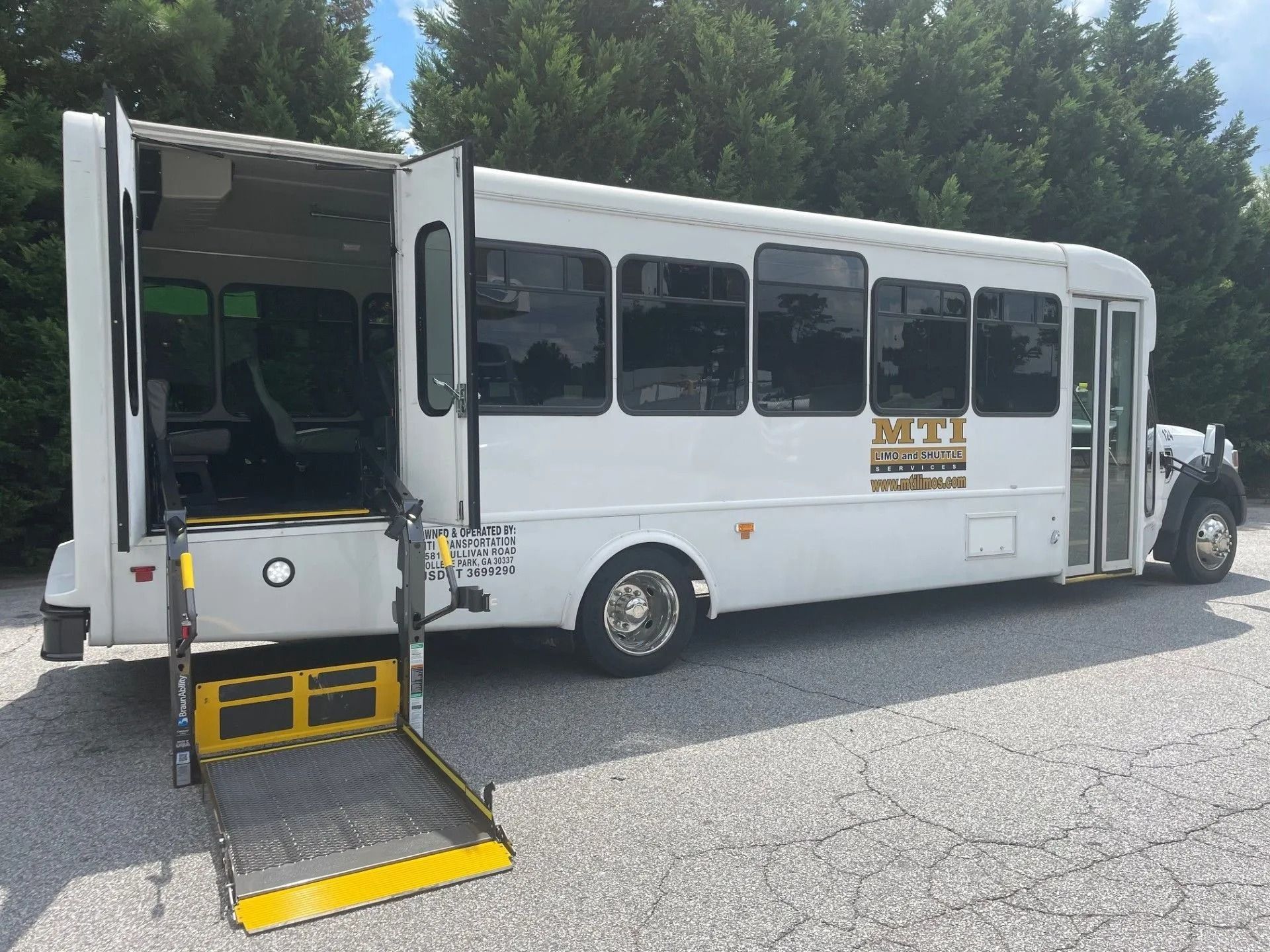 A white shuttle bus with a deployed wheelchair lift parked on a gravel lot in front of trees.