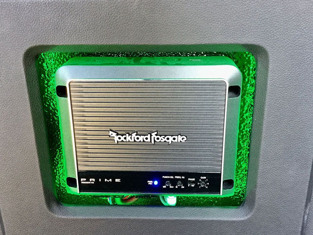Car audio amplifier with green LED lighting, mounted in a vehicle. The amplifier is silver with a Rockford Fosgate logo.
