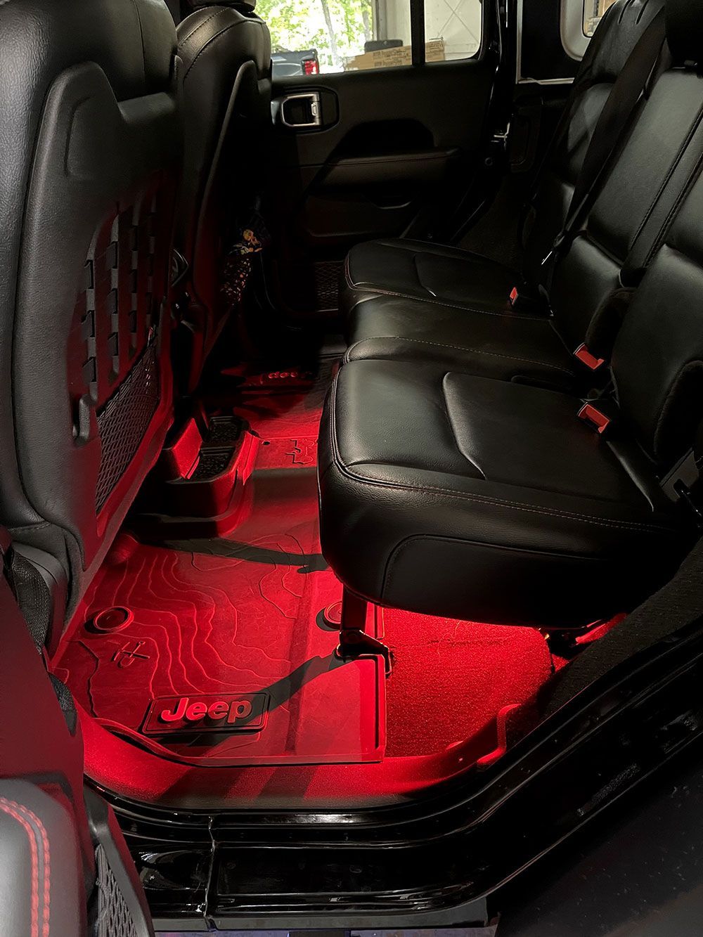 Interior view of a black Jeep with red underglow lighting. Black leather seats, patterned floor mats, and a dark cabin.