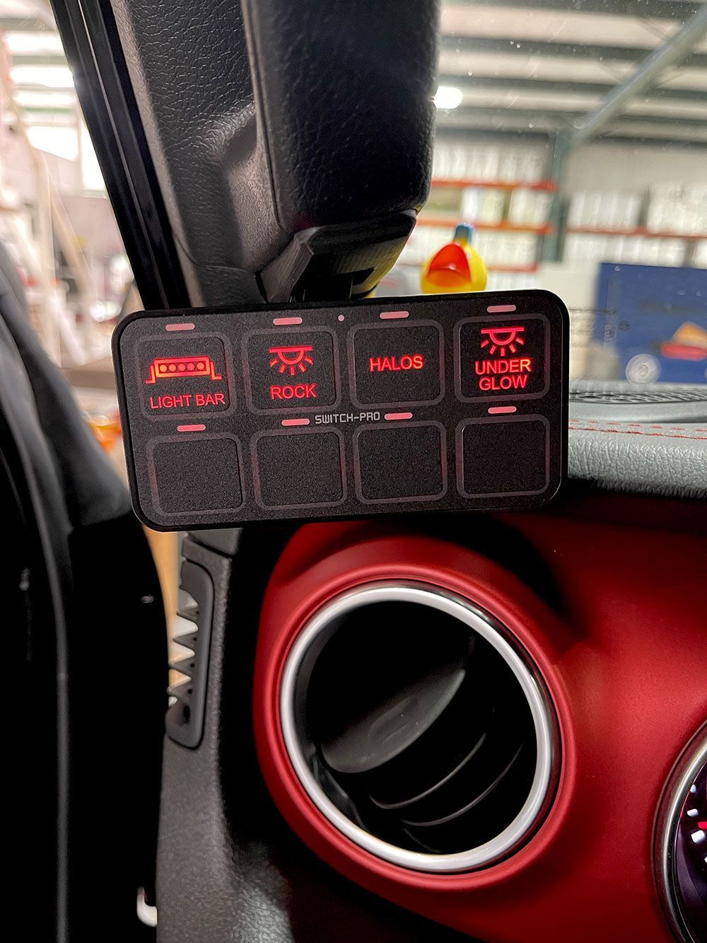 Black switch panel with red illuminated buttons installed in a red-accented car dashboard.