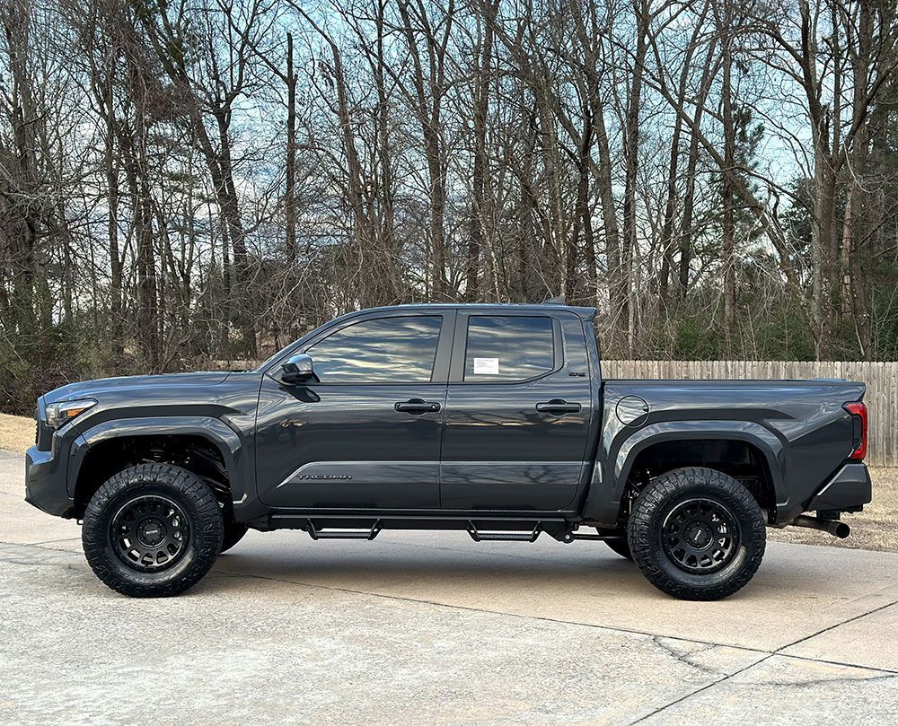 Dark gray pickup truck with black wheels and side steps parked outside, trees in the background.