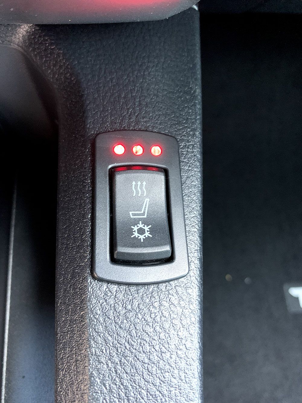 Black car seat heater switch with three red lights illuminated, showing three heat settings.