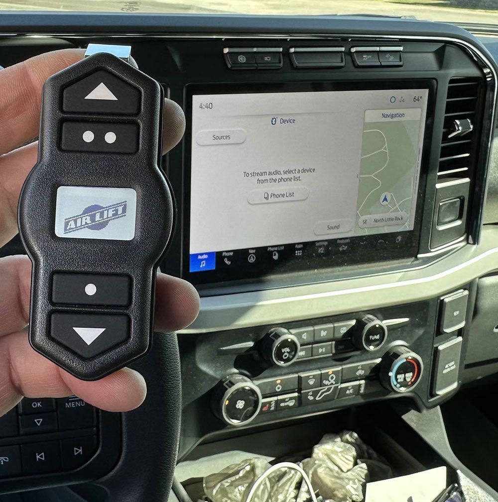 Hand holding an Air Lift remote control near a vehicle's dashboard with a navigation screen visible.