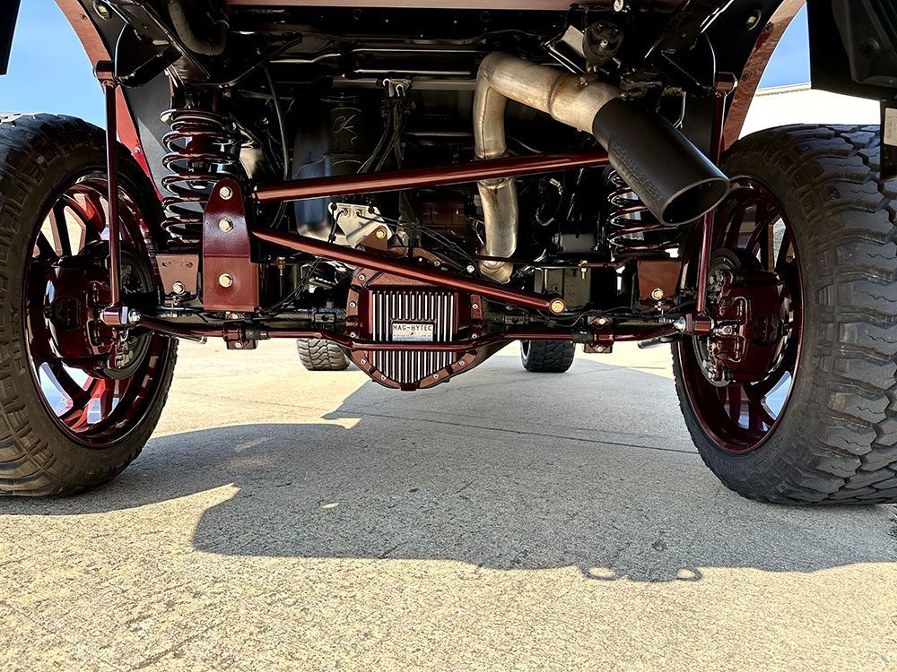 Red truck's lifted rear axle, exhaust, and suspension with large tires visible against a sunny ground.
