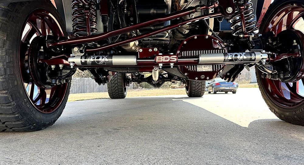 A lifted truck's undercarriage with burgundy suspension, axle, and wheels, parked on a gray surface.