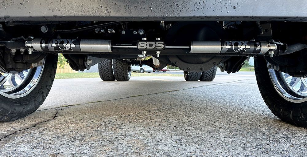 Undercarriage of a truck with a black BDS steering stabilizer system, showing axles and wheels on asphalt.