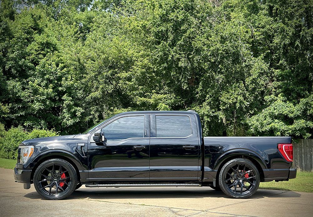 Black Ford F-150 truck with black rims and red brake calipers parked on a paved area, green trees in background.