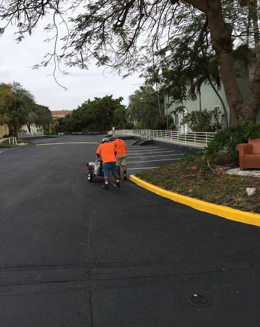 Two workers in orange shirts push a pavement marking machine on a newly paved road. Yellow curb on right.