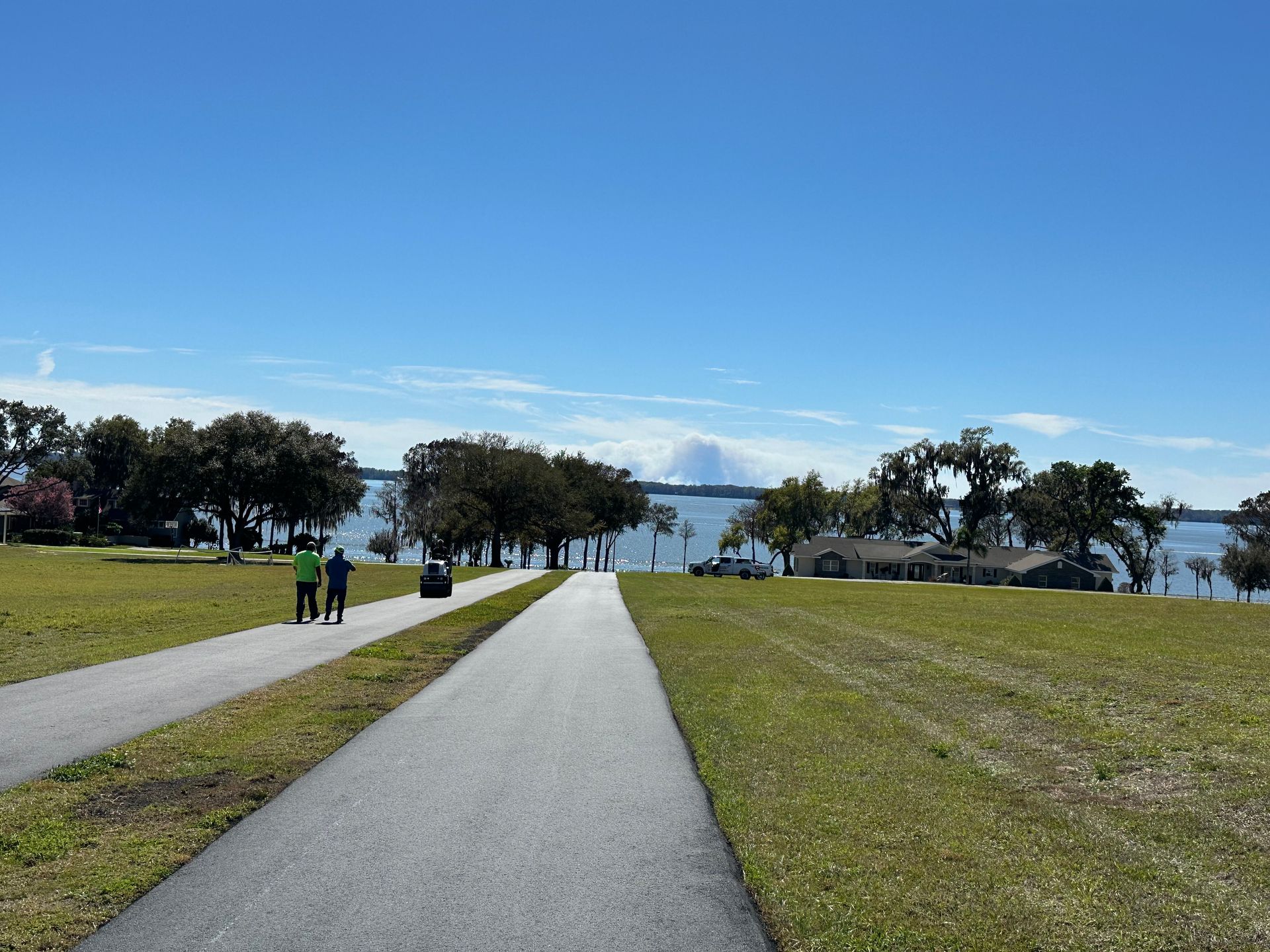 Paved path through grassy area towards lake with trees and blue sky. Two people walk.