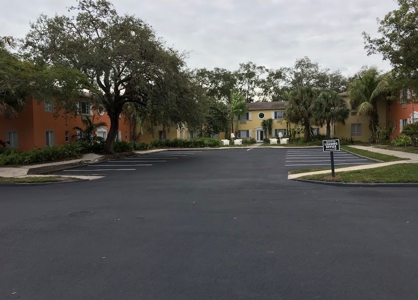 Empty asphalt parking lot in front of multi-colored apartments with trees under an overcast sky.