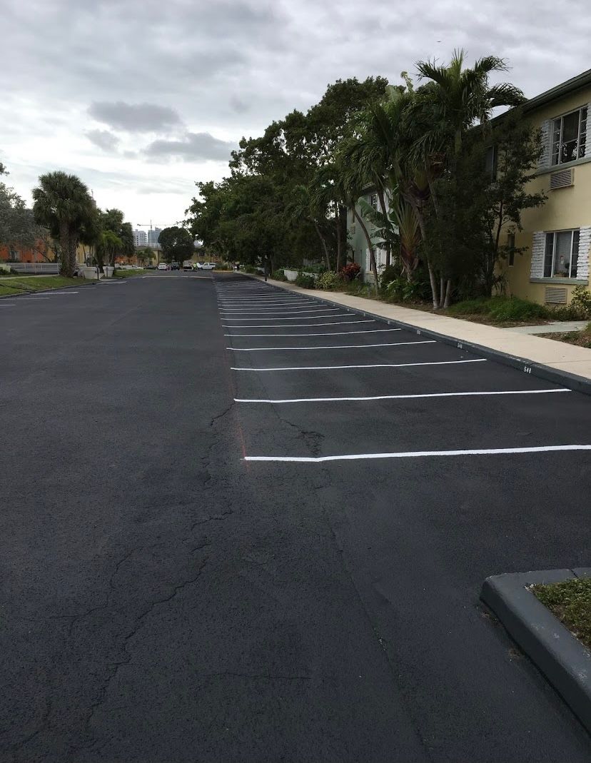 Black asphalt parking lot with white painted parking space lines, next to a building and trees.