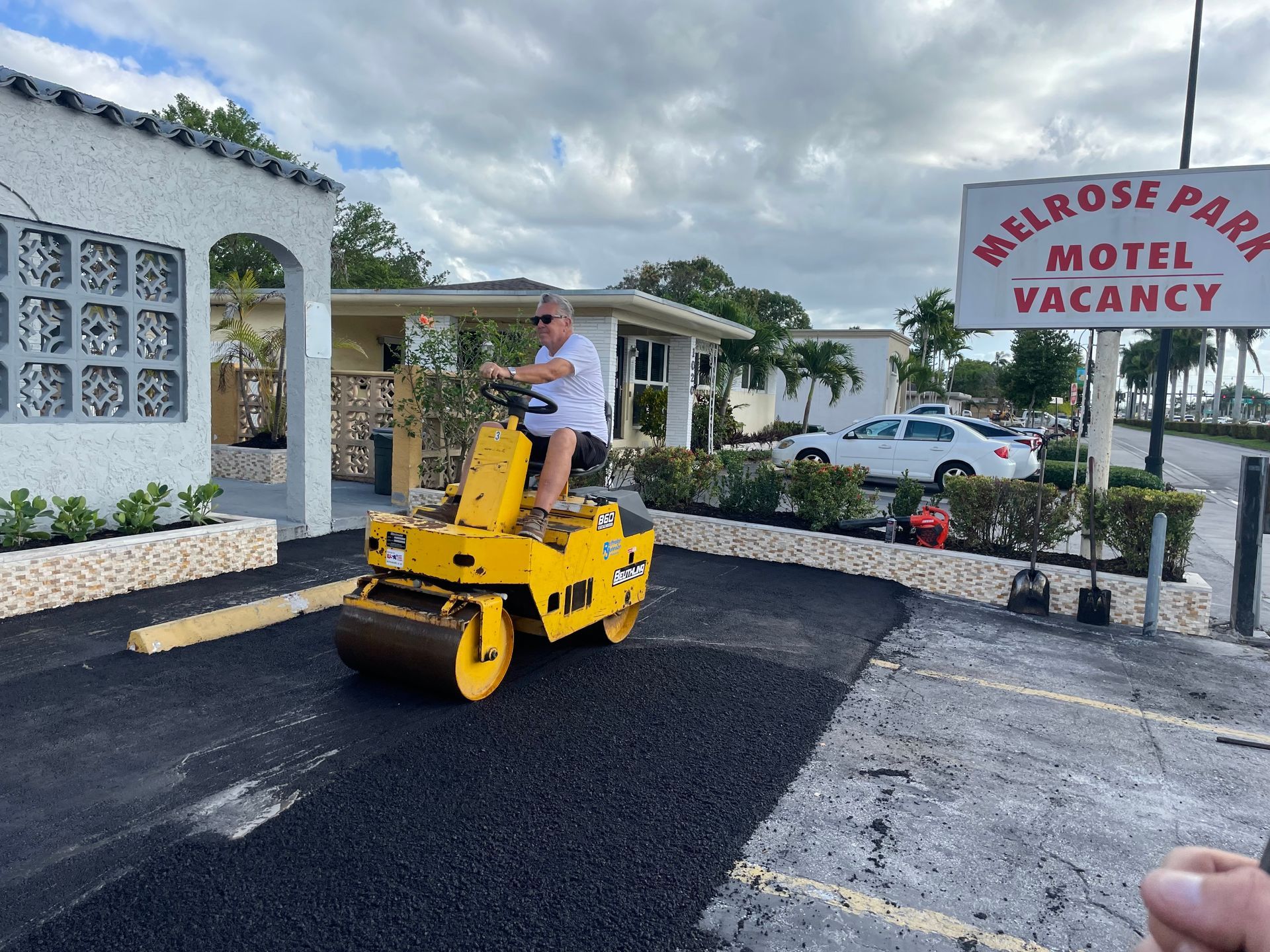 Man operating a yellow road roller, paving asphalt near the Melrose Park Motel sign.