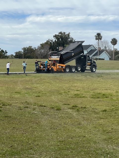Dump truck offloading asphalt into a paver on a grassy area, with two people supervising.