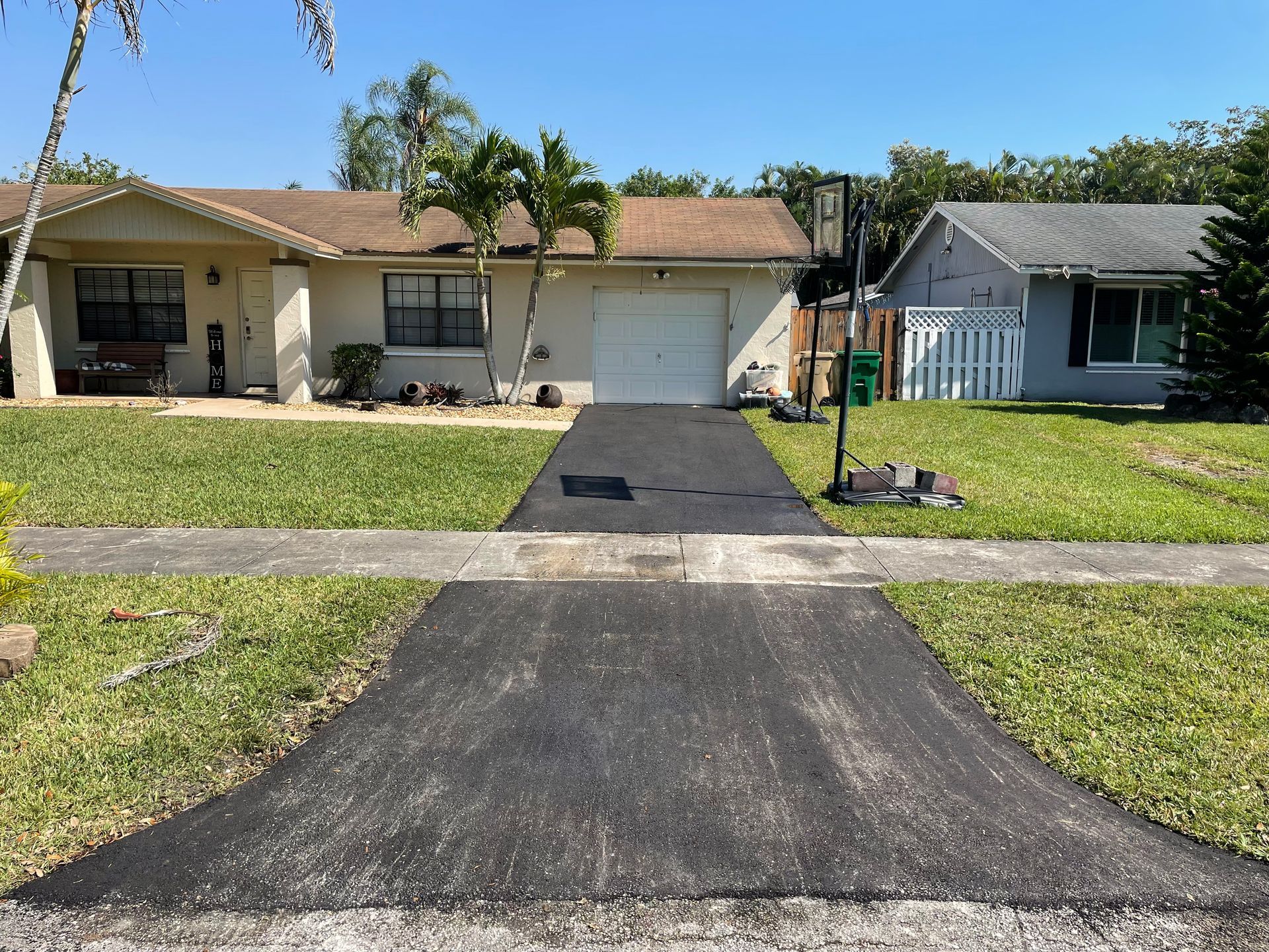 Newly paved black asphalt driveway leading to a beige house with a white garage door, set in a sunny, residential area.