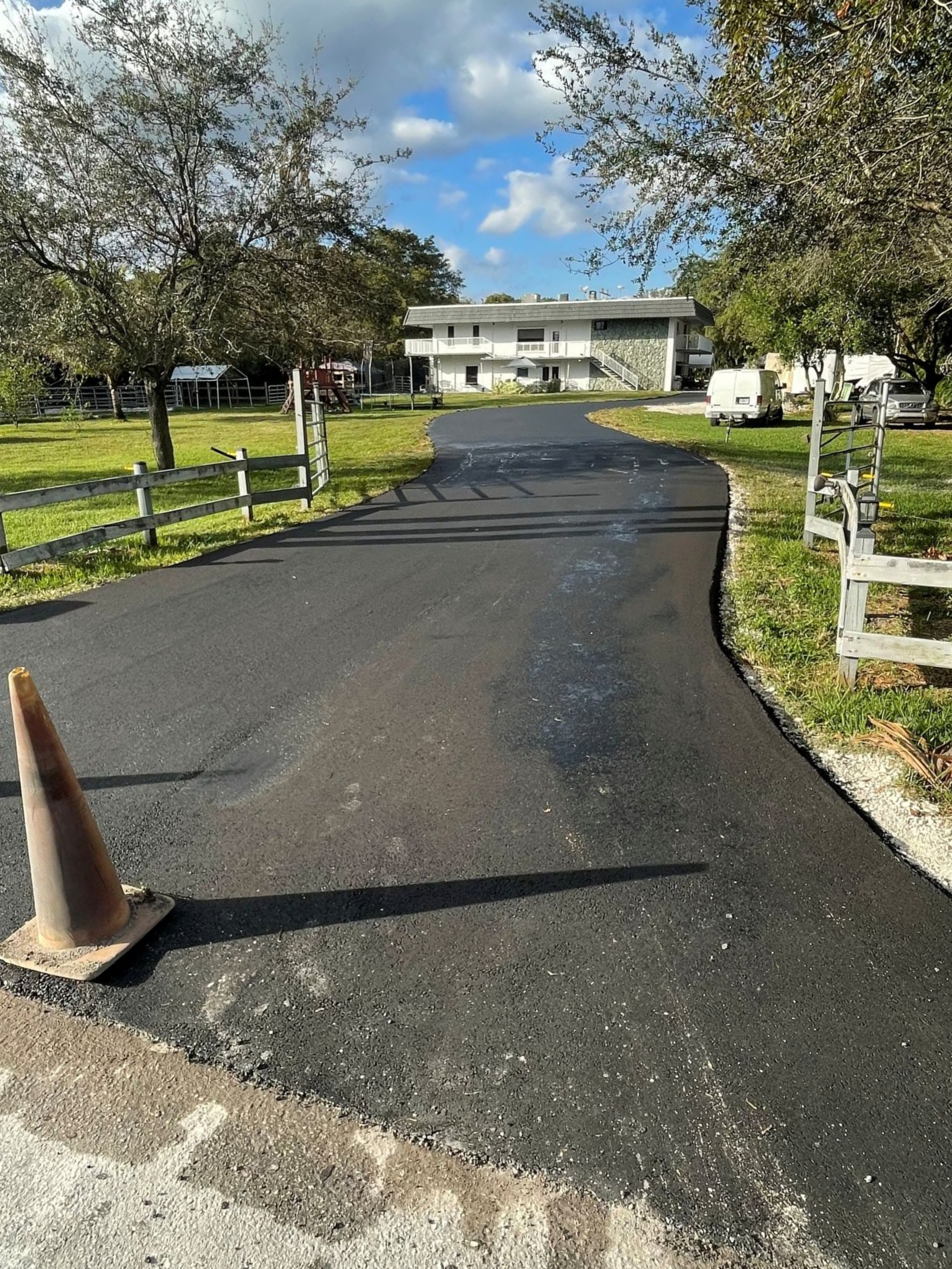 Freshly paved asphalt driveway with orange traffic cones on the side, white fence, green grass, and trees.