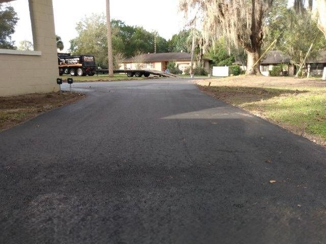 Newly paved black asphalt driveway leading towards houses, with grassy areas on both sides.