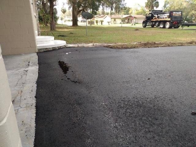 Freshly paved asphalt driveway next to a white house with a utility truck in the background.