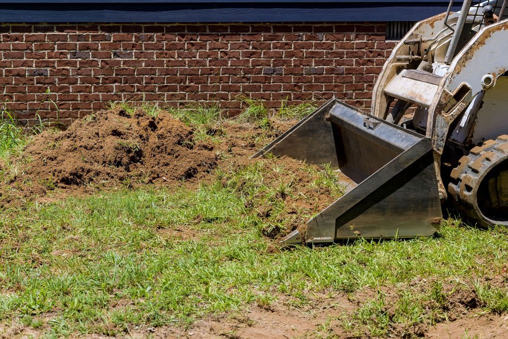 Skid steer loader scoops soil from a grassy area in front of a brick building.
