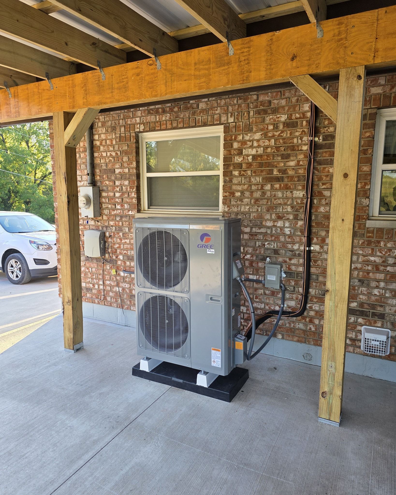An outdoor air conditioning unit under a wooden canopy attached to a brick building.