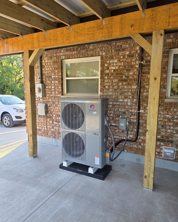 An outdoor air conditioning unit under a wooden canopy attached to a brick building.