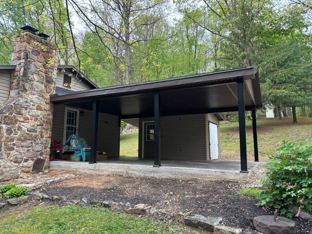 Stone cottage with a covered carport and teal chairs, set among trees in a wooded yard.