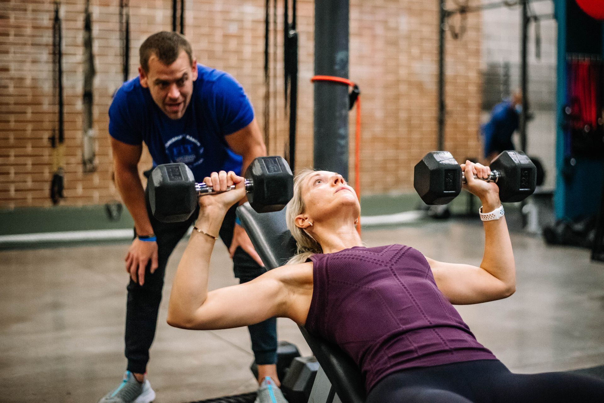 A man is watching a woman lift dumbbells on a bench in a gym.