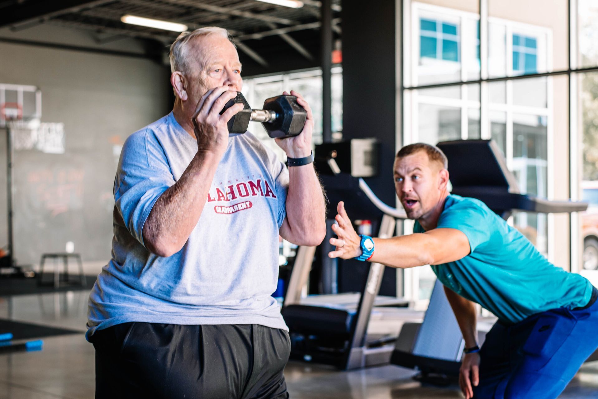 A man is helping an older man lift a dumbbell in a gym.