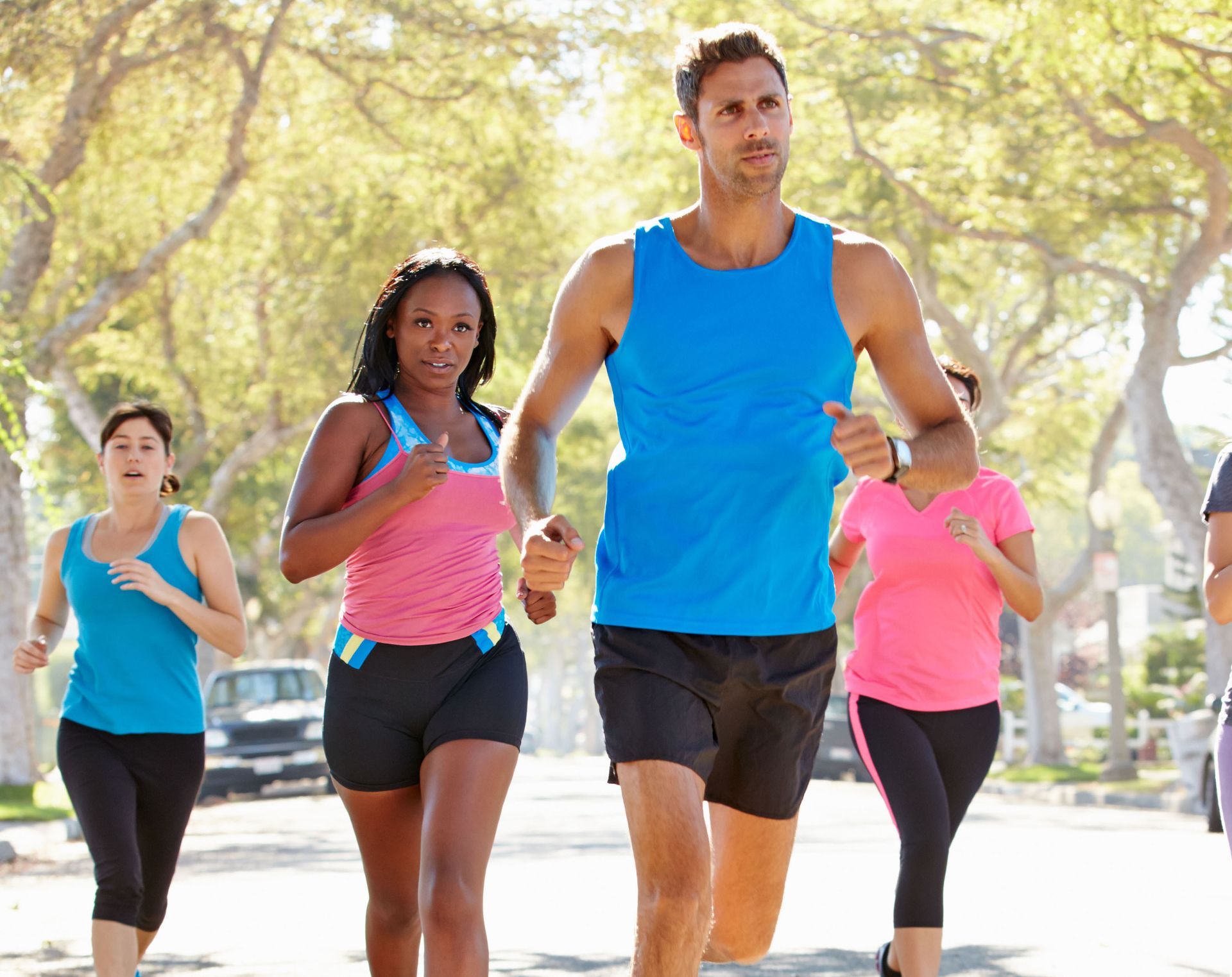 Group of people running down a sunny street; man in blue tank top leads, others in activewear.