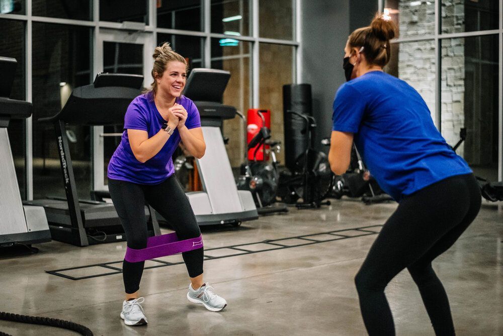 Two women are doing squats with resistance bands in a gym.