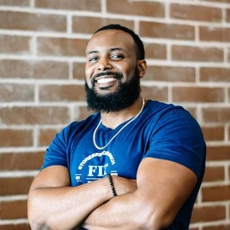 A man with a beard is standing in front of a brick wall with his arms crossed.