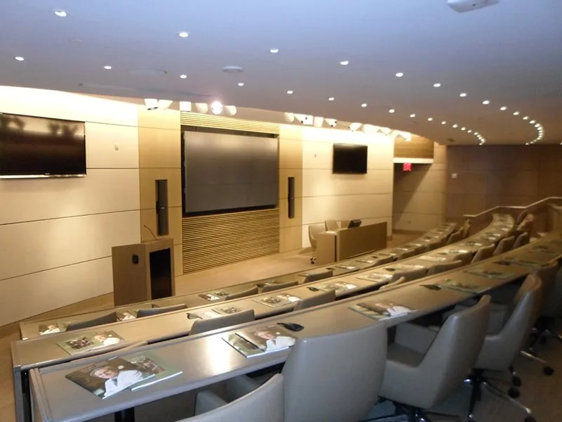 A tiered conference room with rows of tables, gray chairs, and a large projection screen at the front.