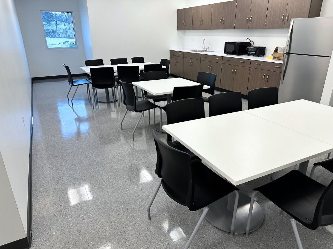 Breakroom with rows of white square tables, black chairs, kitchen cabinets, and a refrigerator against a white wall.