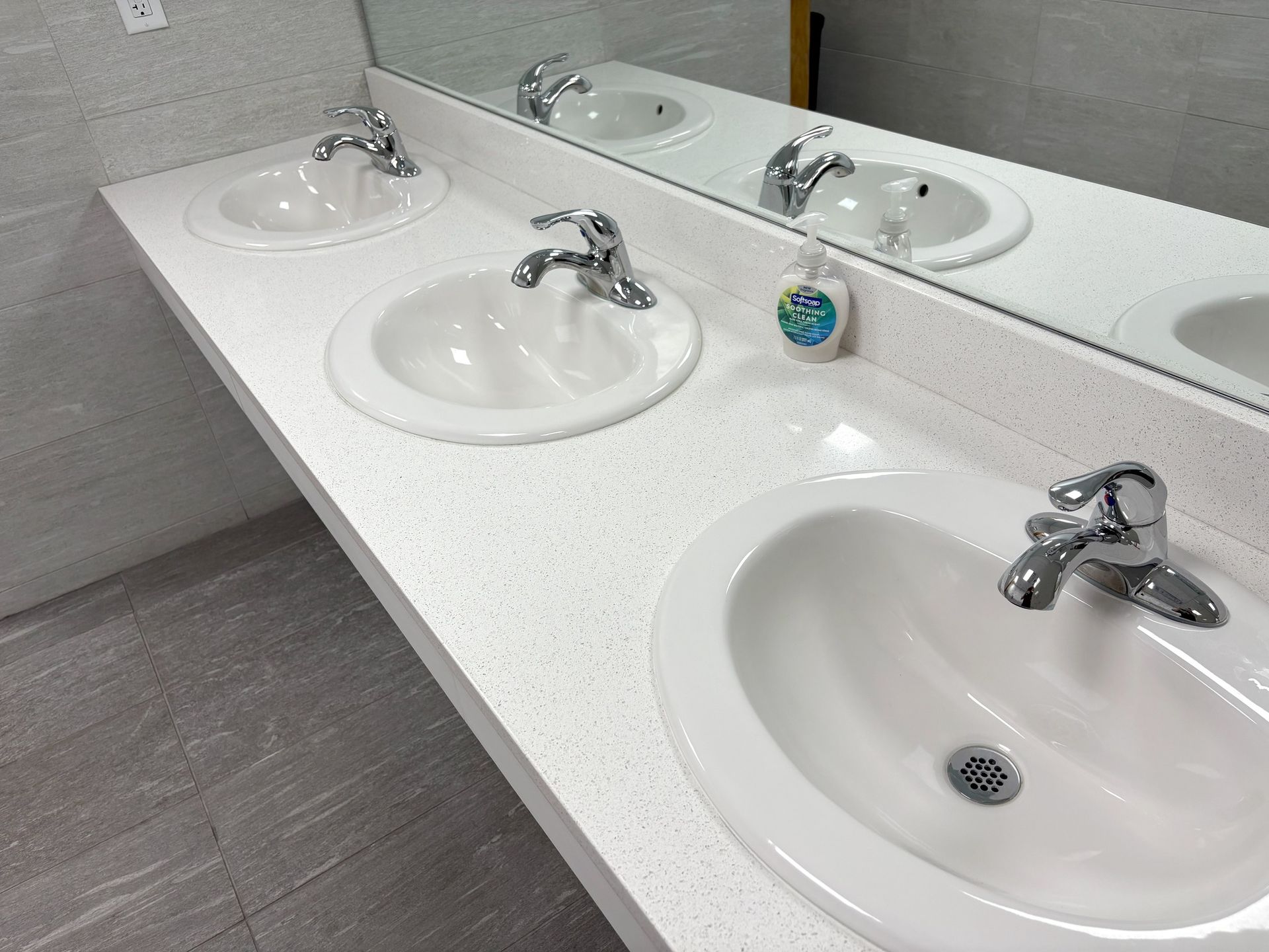 Three white sinks with chrome faucets on a speckled countertop in a public restroom with a mirror.
