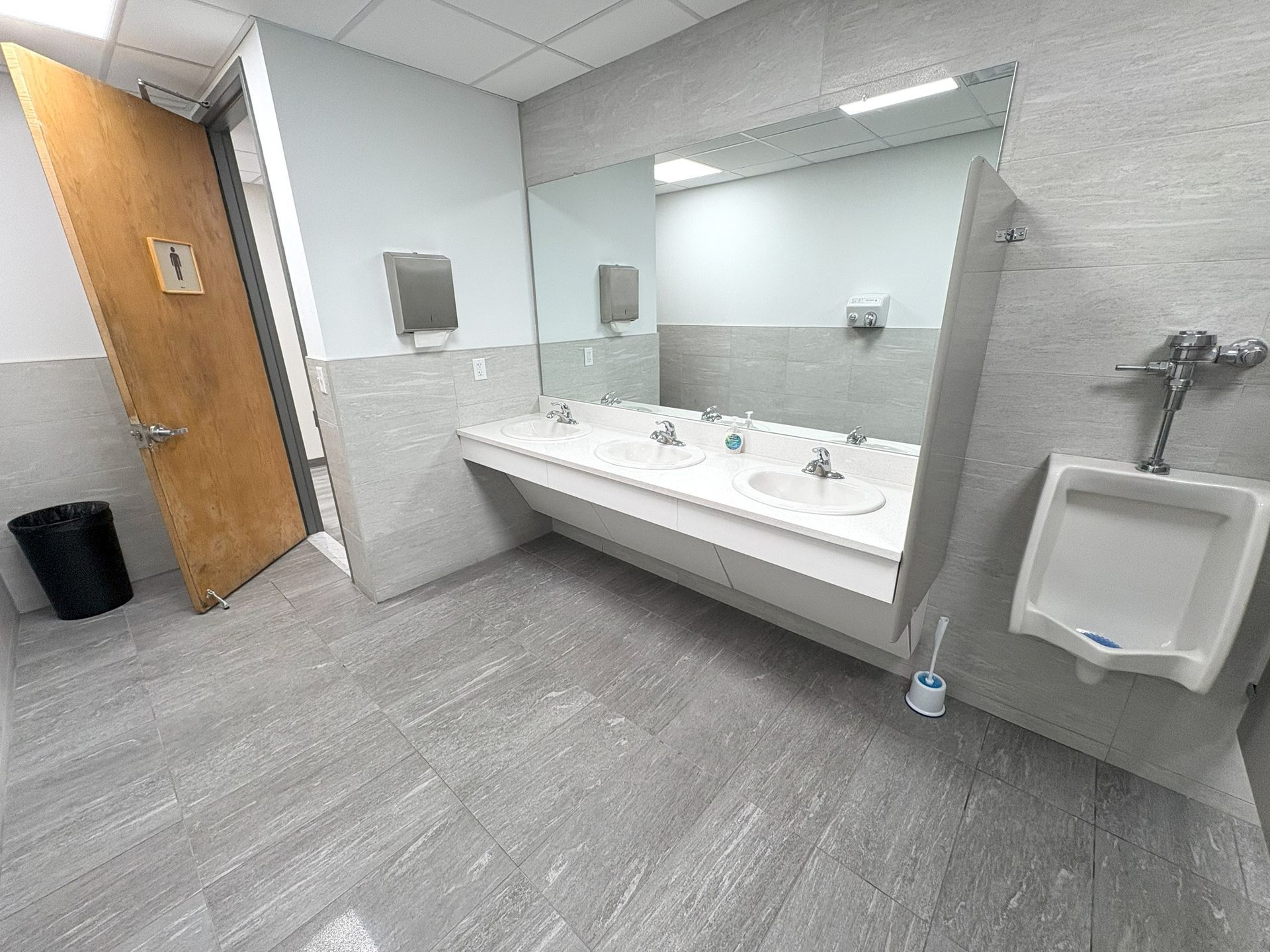Public restroom with sinks, urinals, and a door. Gray and white color scheme.
