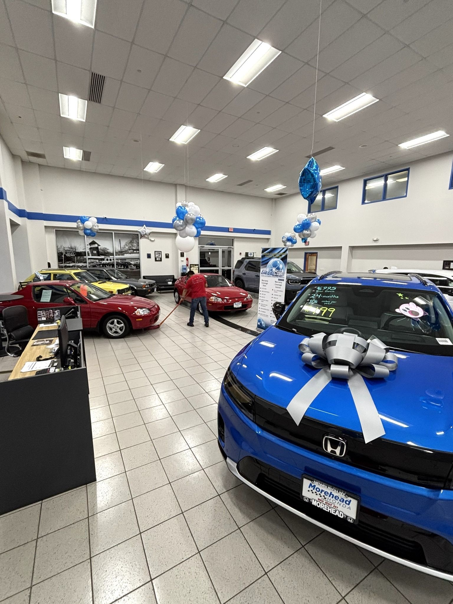 Blue car in a showroom with a large bow, balloons, and people.