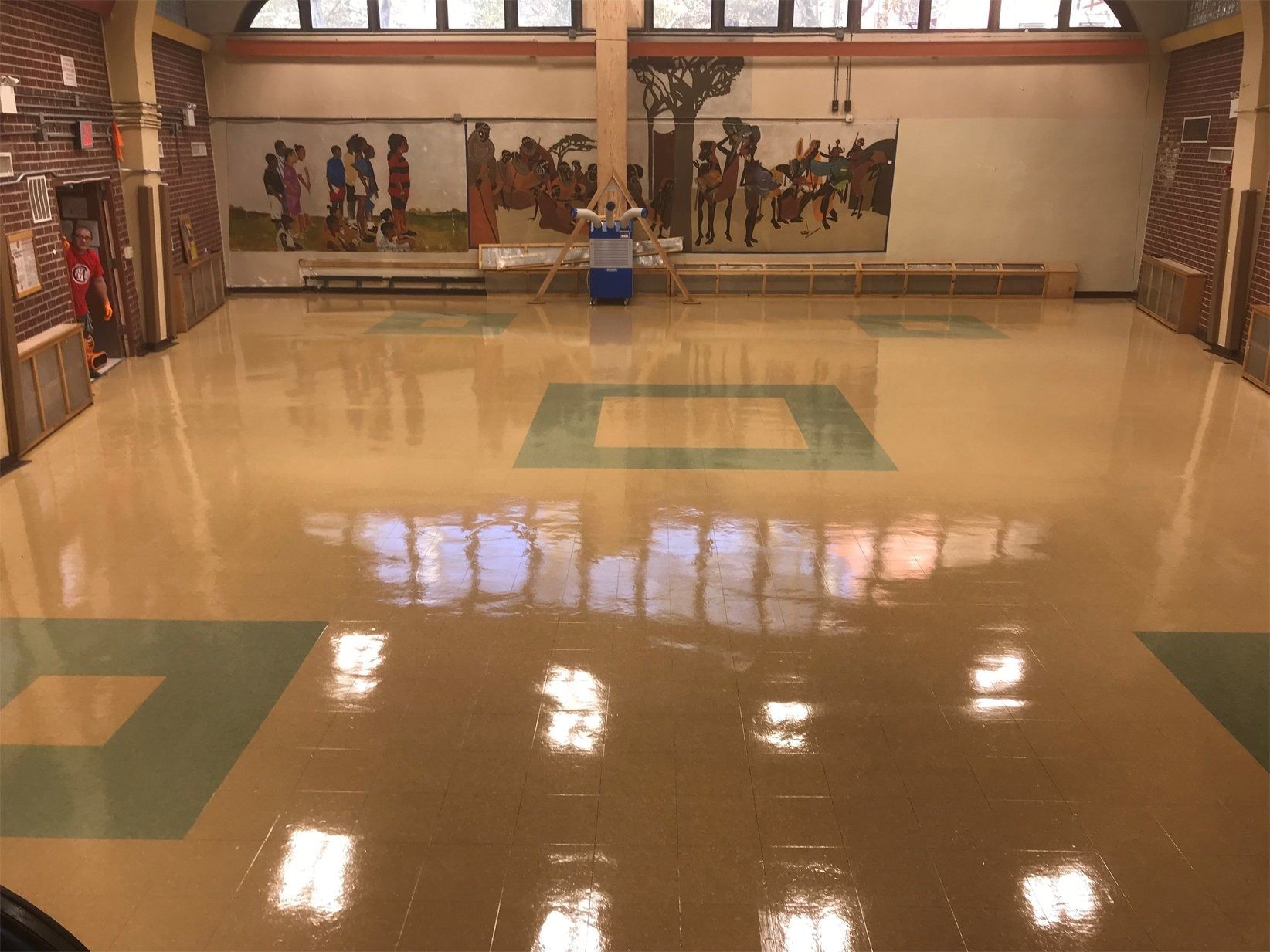 A polished, shiny school hall with tan and green floor tiles, featuring murals on the back walls and a central pillar.