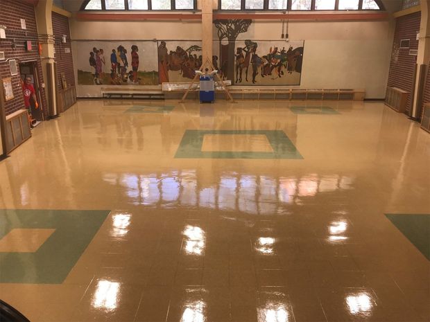 An indoor community space with a glossy tan floor featuring green square accents, a murals-lined back wall, and windows.