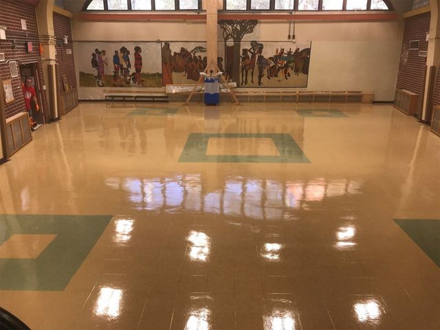 An indoor community space with a glossy tan floor featuring green square accents, a murals-lined back wall, and windows.