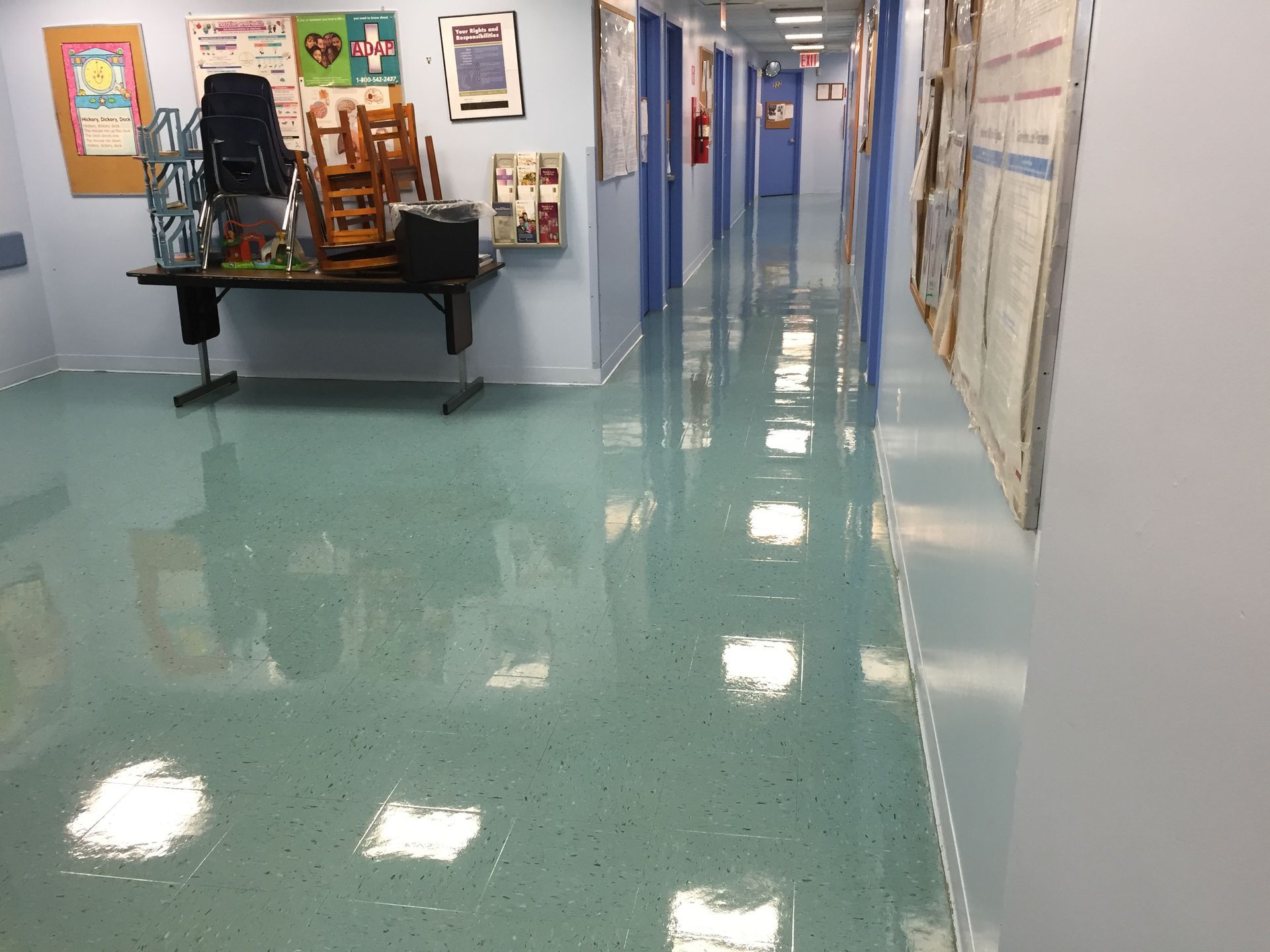 Hallway with glossy turquoise floor and light blue walls. Posters, tables, and doors line the sides.
