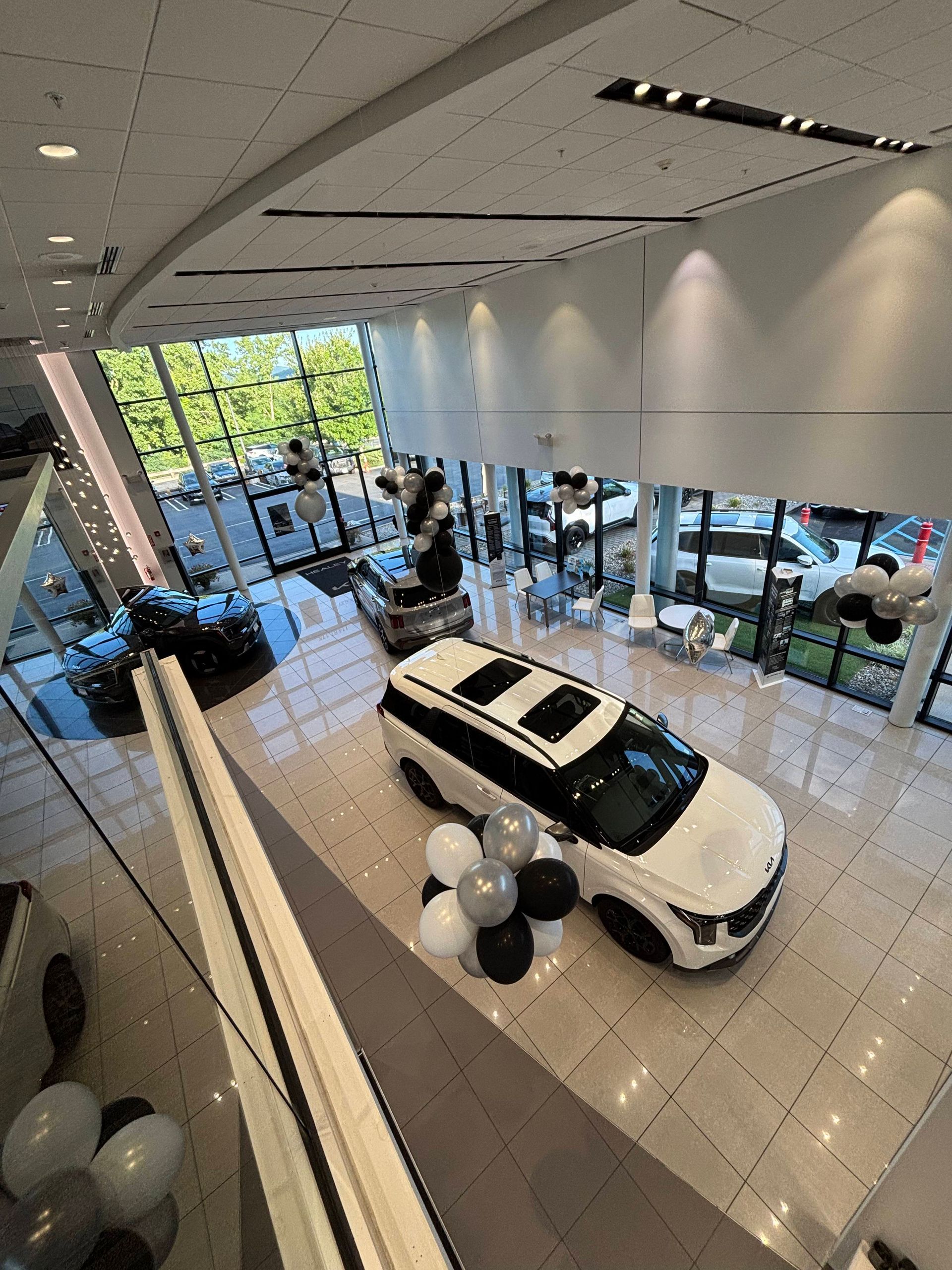 White SUV inside a car dealership showroom with balloons and large windows overlooking exterior.