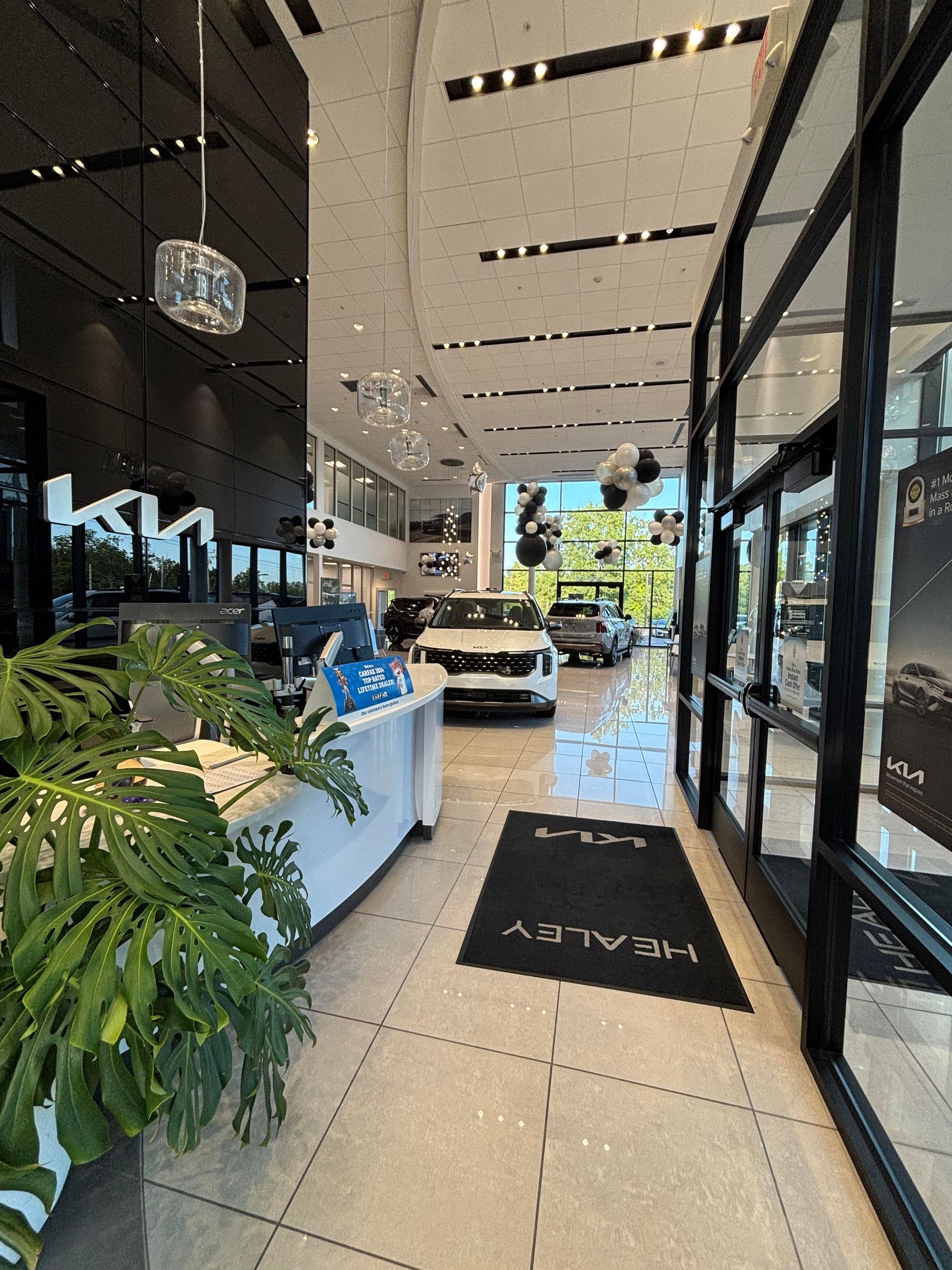 Interior of a car dealership. White and black decor, vehicles on display, reception desk with a plant.