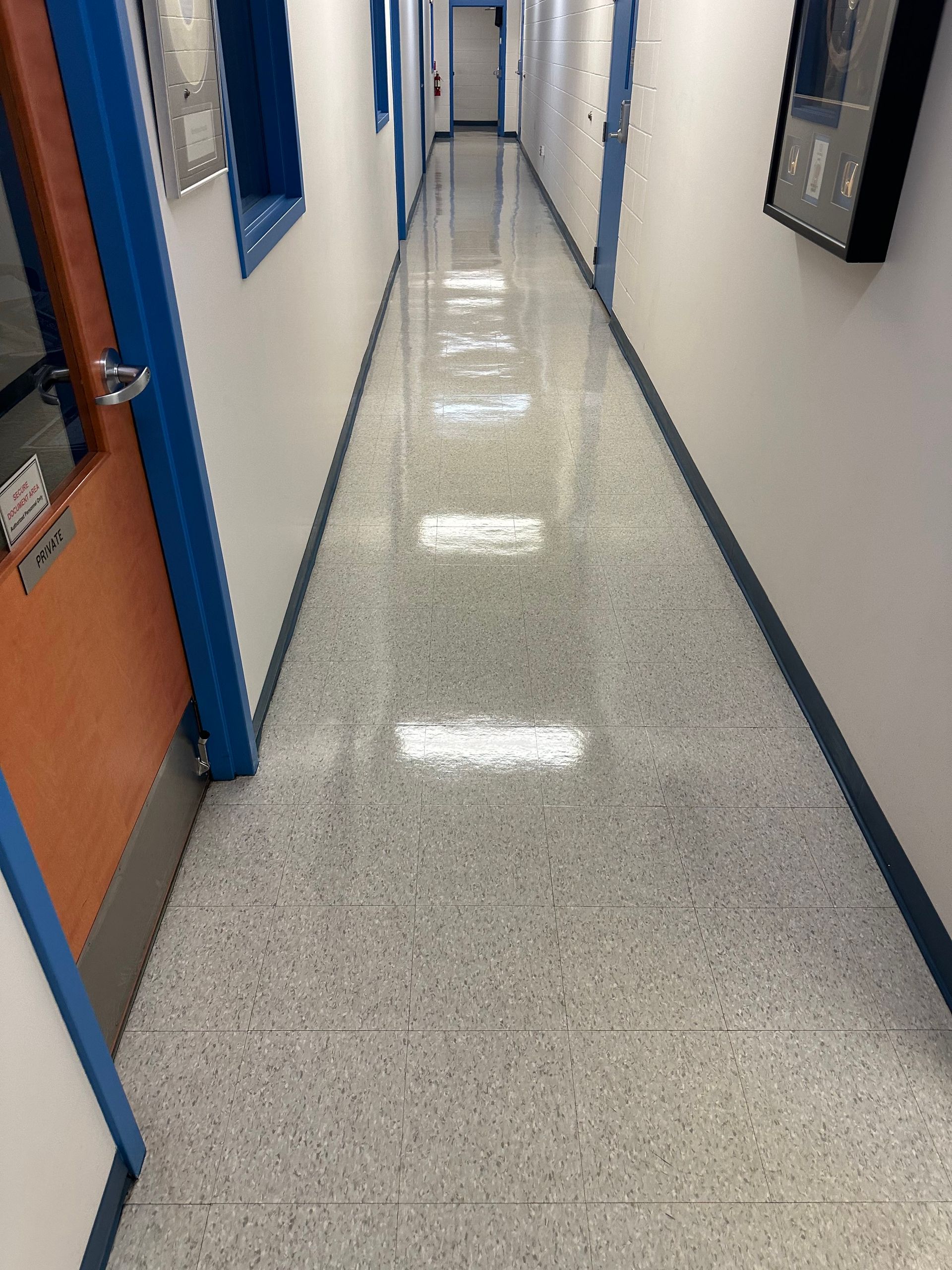 Long, brightly lit institutional hallway with speckled flooring, doors on the left, and a frame on the right.