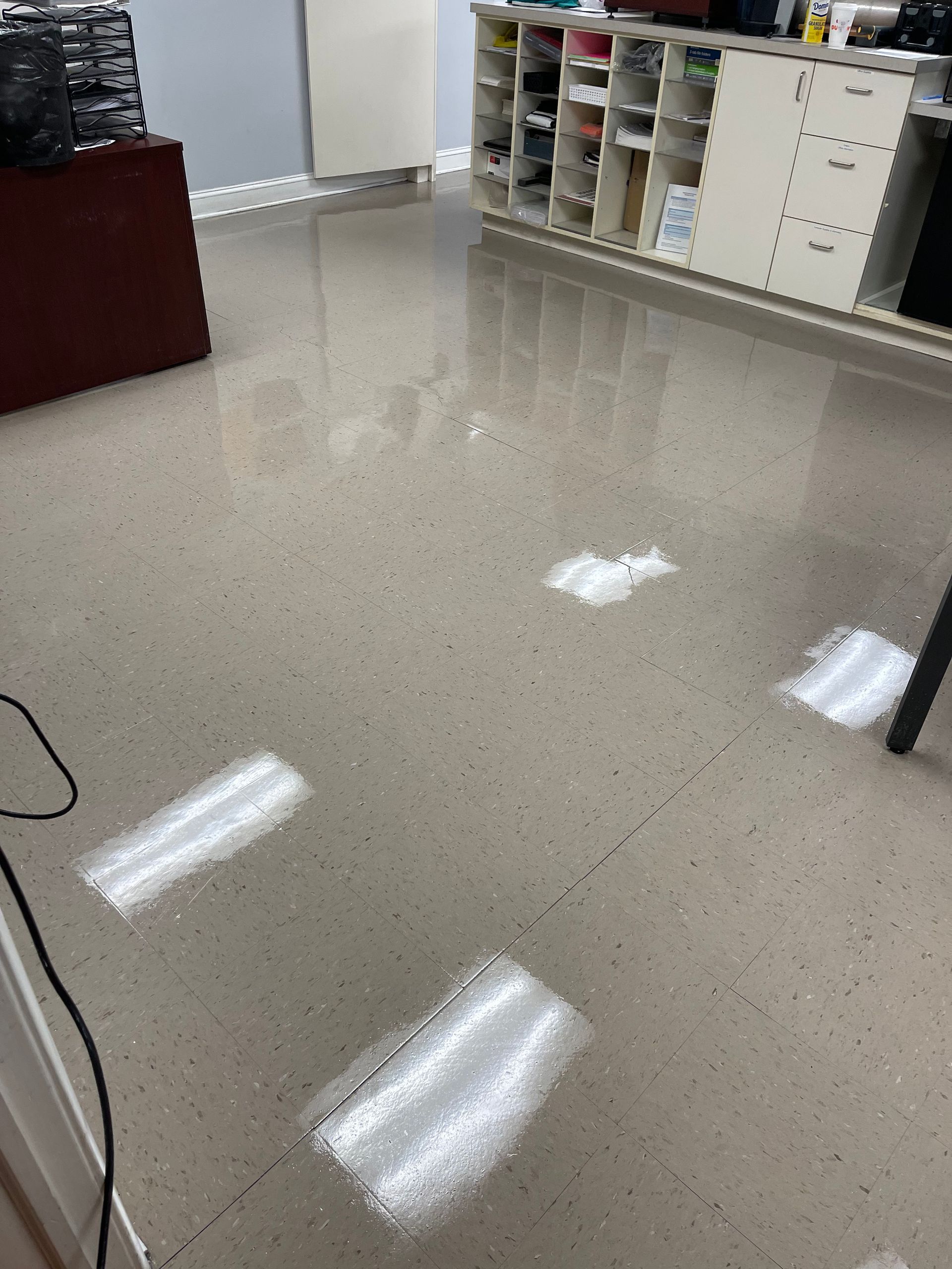A high-angle view of a reflective, beige-speckled office floor with bright white light reflections from overhead fixtures.