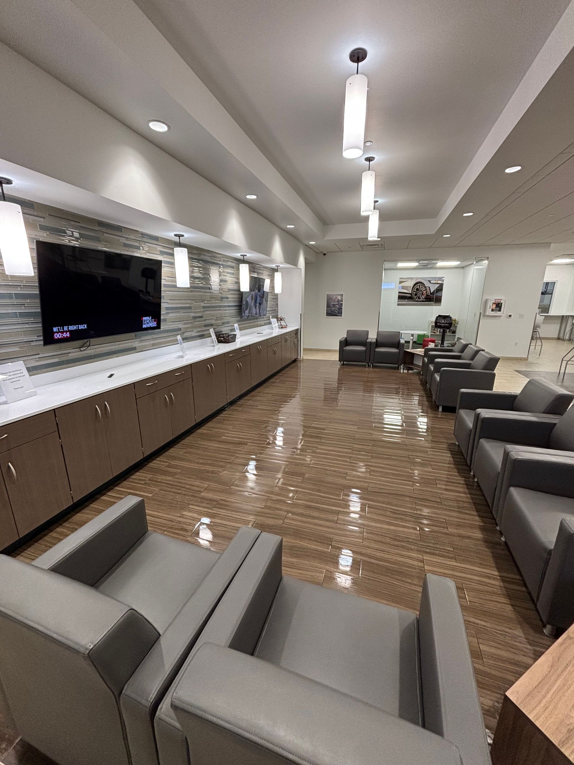 Modern waiting room with grey armchairs, long cabinet with TV, and pendant lights.