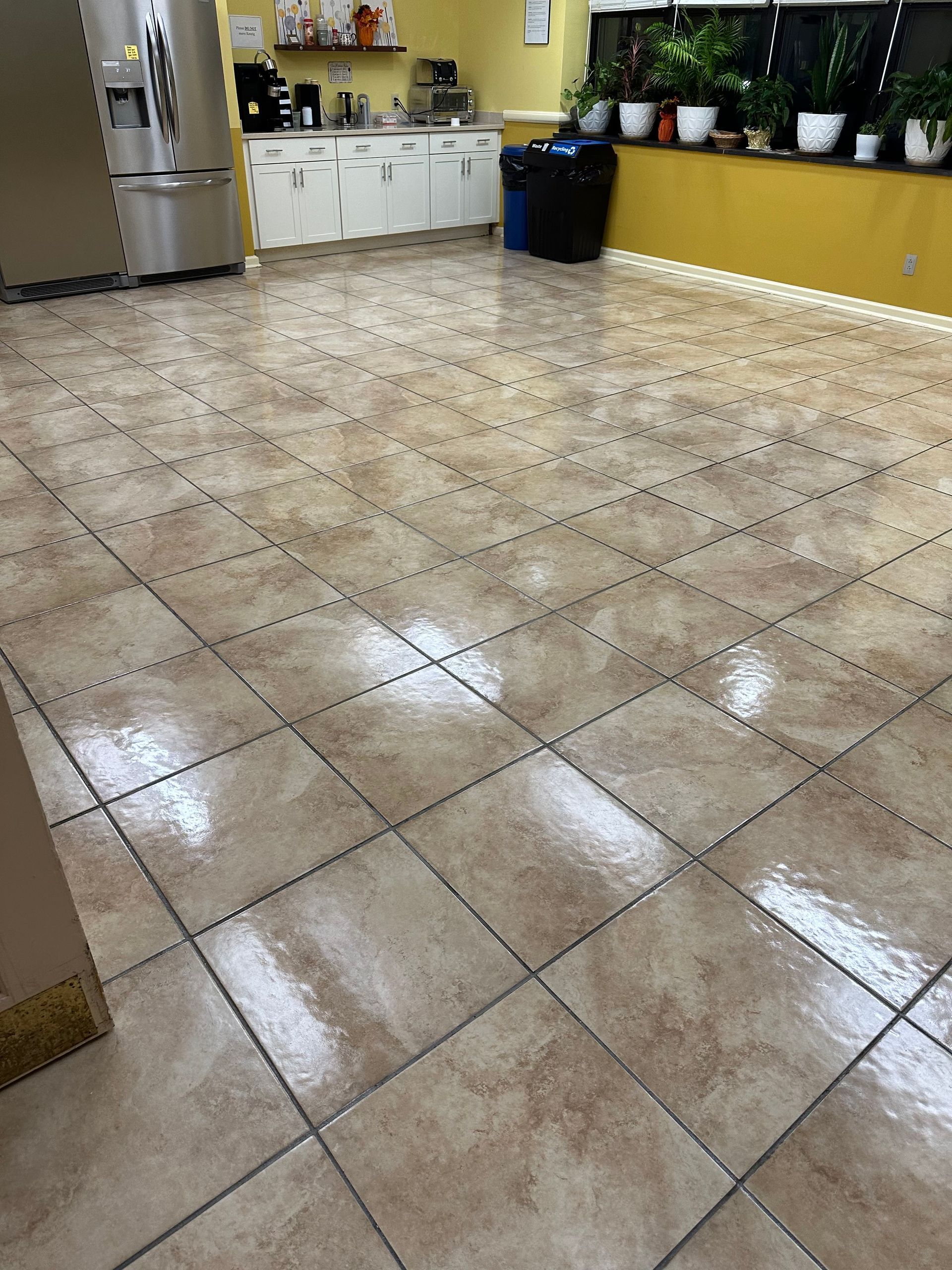 A polished, light-brown tiled floor in a kitchen with white cabinetry, a refrigerator, and potted plants on a ledge.