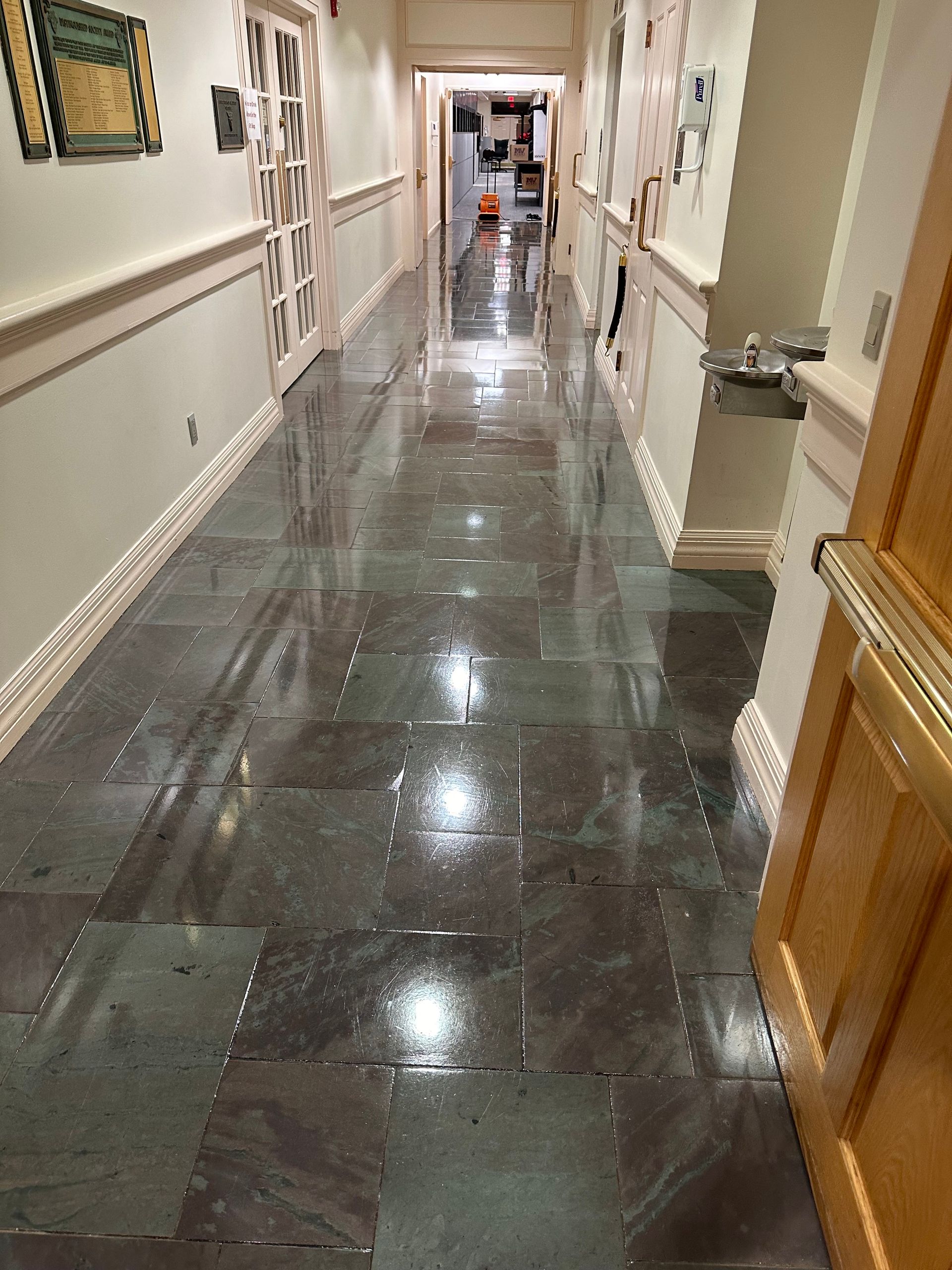 A long, narrow indoor hallway with glossy, dark-toned stone floor tiles reflecting overhead lighting.